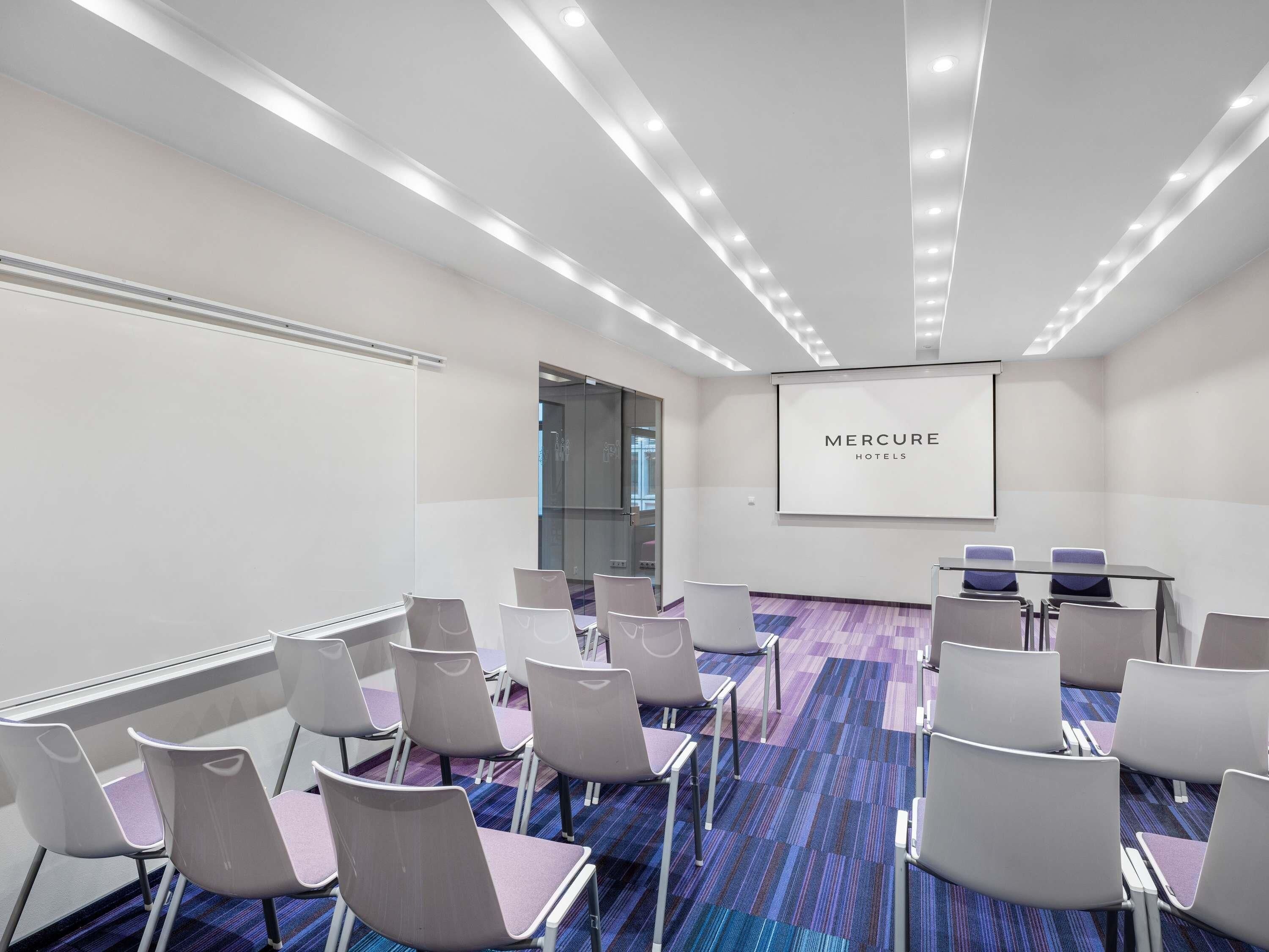 A conference room with a glass partition, a screen displaying 'MERCURE HOTELS', and rows of chairs with a patterned carpet.