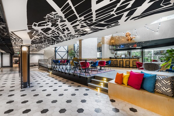 Un vestíbulo moderno con un diseño geométrico en el techo, una zona de bar y un área de descanso con cojines de colores.
