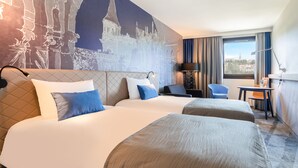 A hotel room with two beds, a desk, a chair, and a large window with a view of a cityscape. - Mercure Budapest Castle Hill (Budapest)