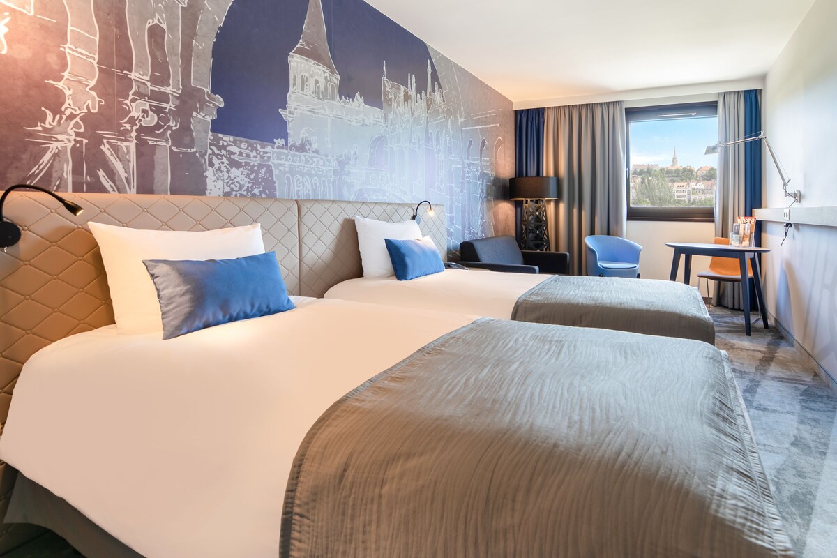 Habitación de hotel con dos camas, un escritorio, una silla y un ventanal con vista a la ciudad.