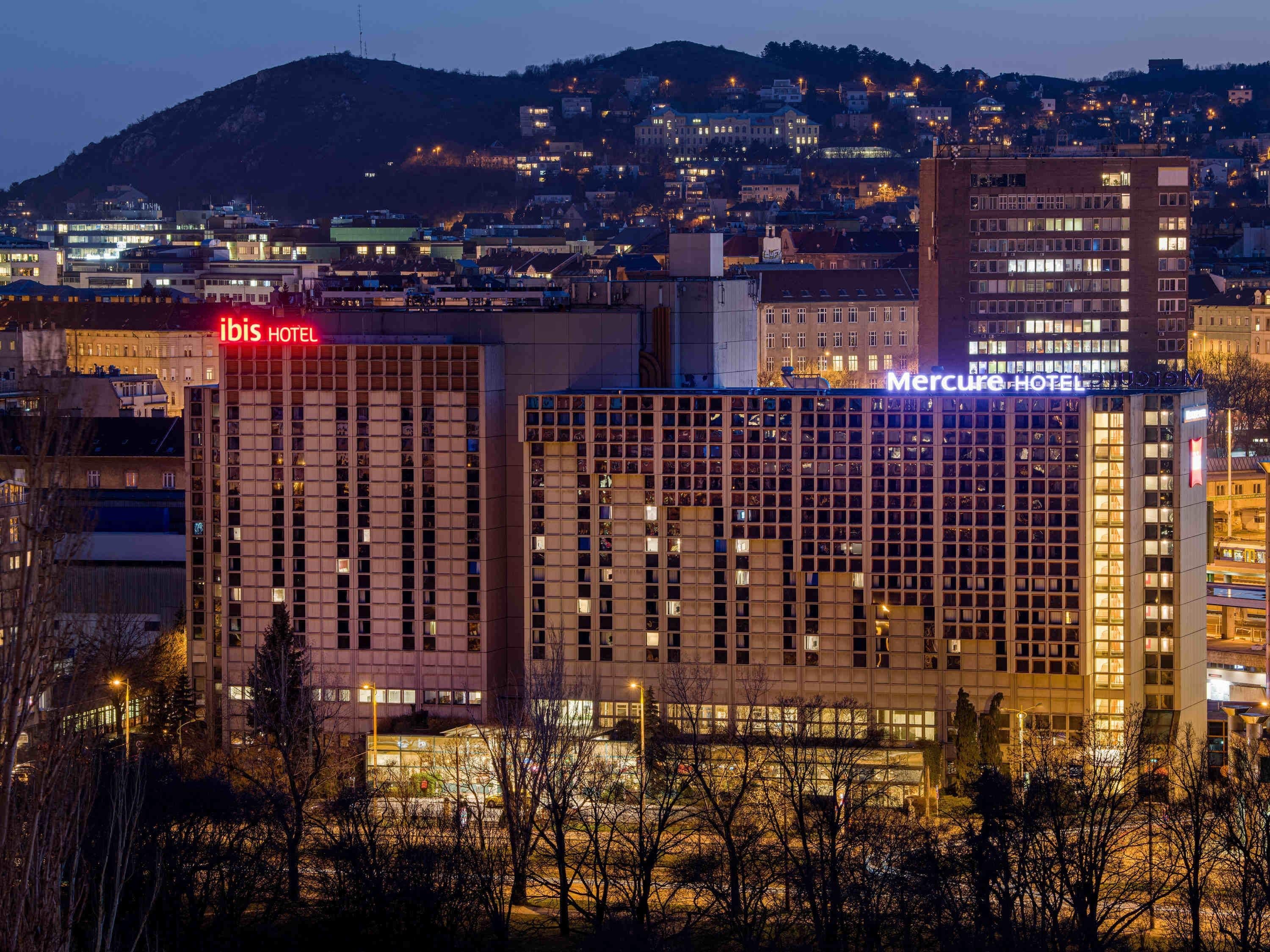 Um hotel com fachada em estilo de grade, uma placa de identificação visível e vista para o horizonte da cidade ao fundo.