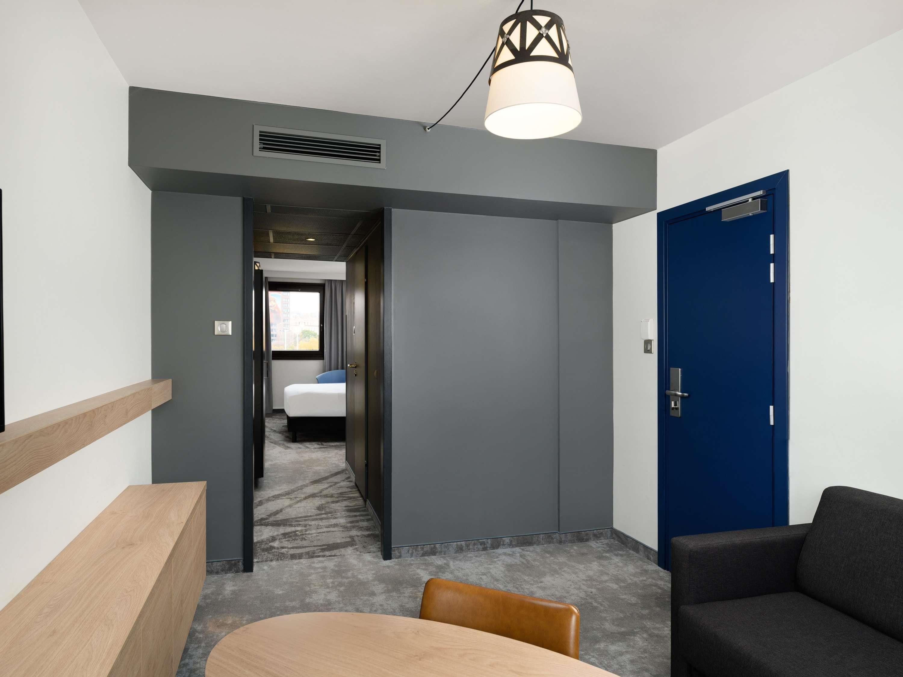 Une chambre d’hôtel avec un lit, un bureau, une chaise, une porte bleue et un couloir menant à une autre chambre.