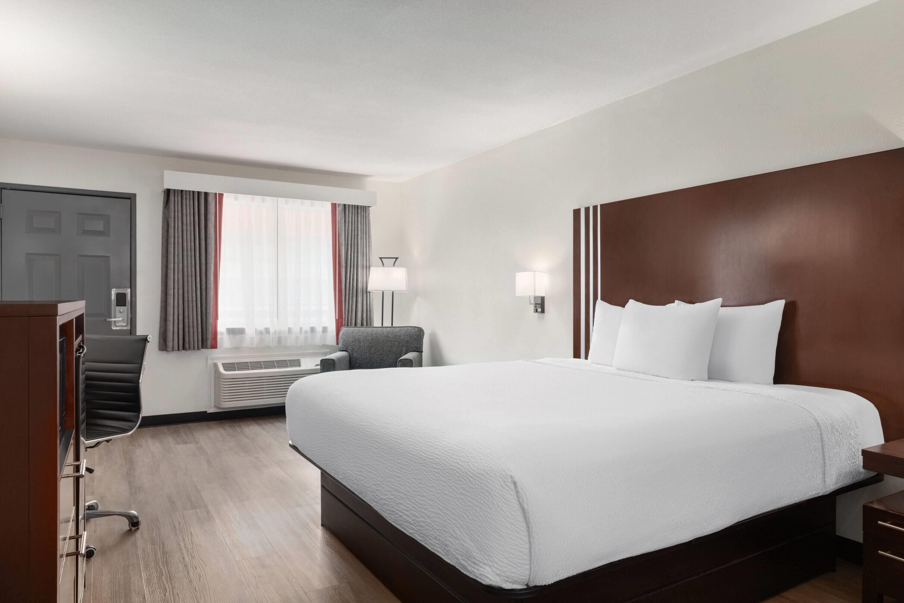 A hotel room with a large bed, a desk, a chair, a window with curtains, and a wall-mounted lamp.