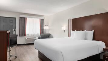 A hotel room with a large bed, a desk, a chair, a window with curtains, and a wall-mounted lamp.