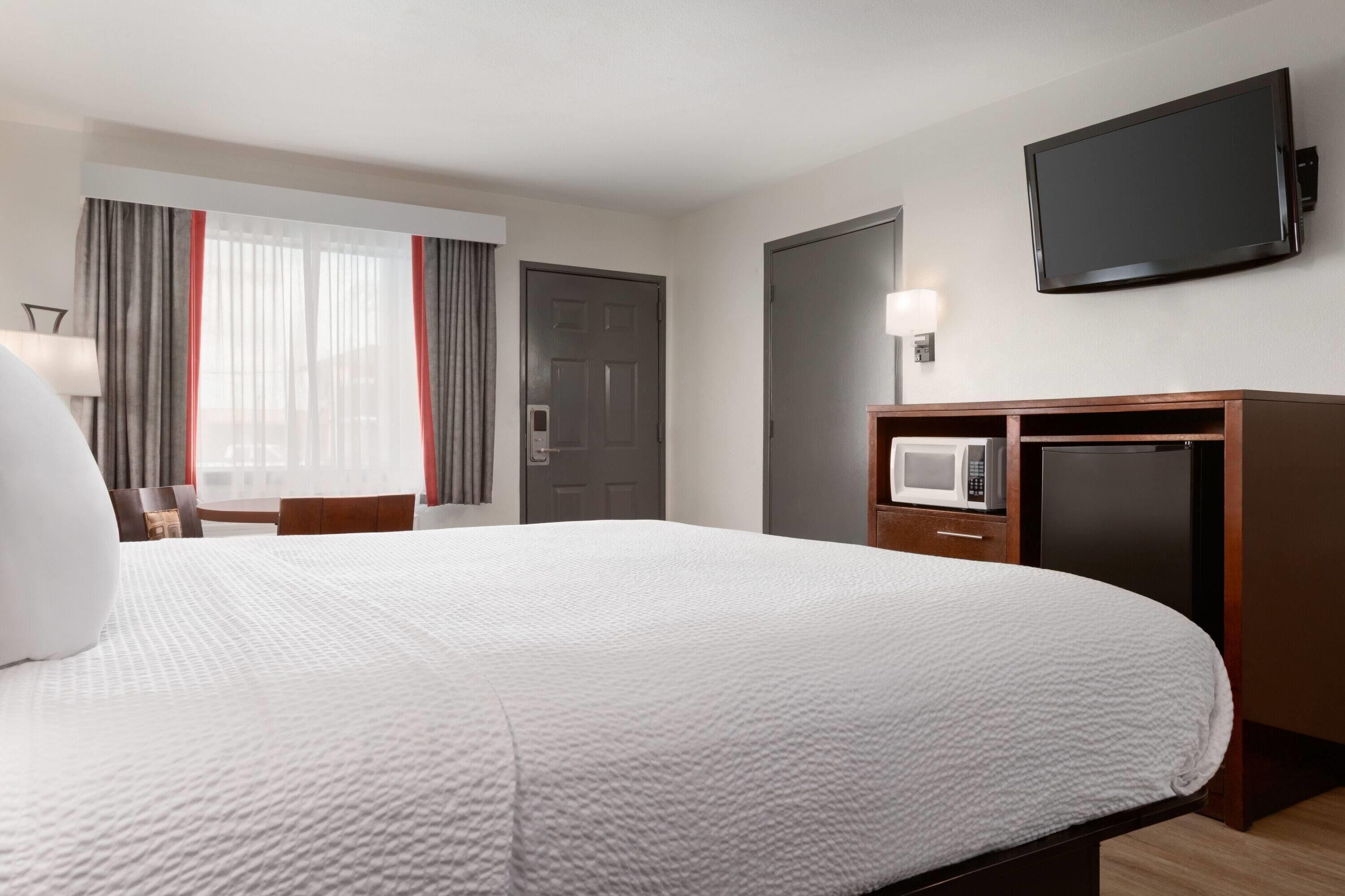 A hotel room with a large bed, a flat-screen TV, a desk with a lamp, a microwave, and a refrigerator.