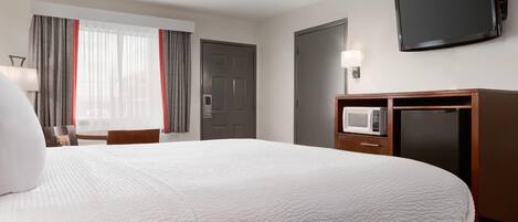 A hotel room with a large bed, a flat-screen TV, a desk with a lamp, a microwave, and a refrigerator.