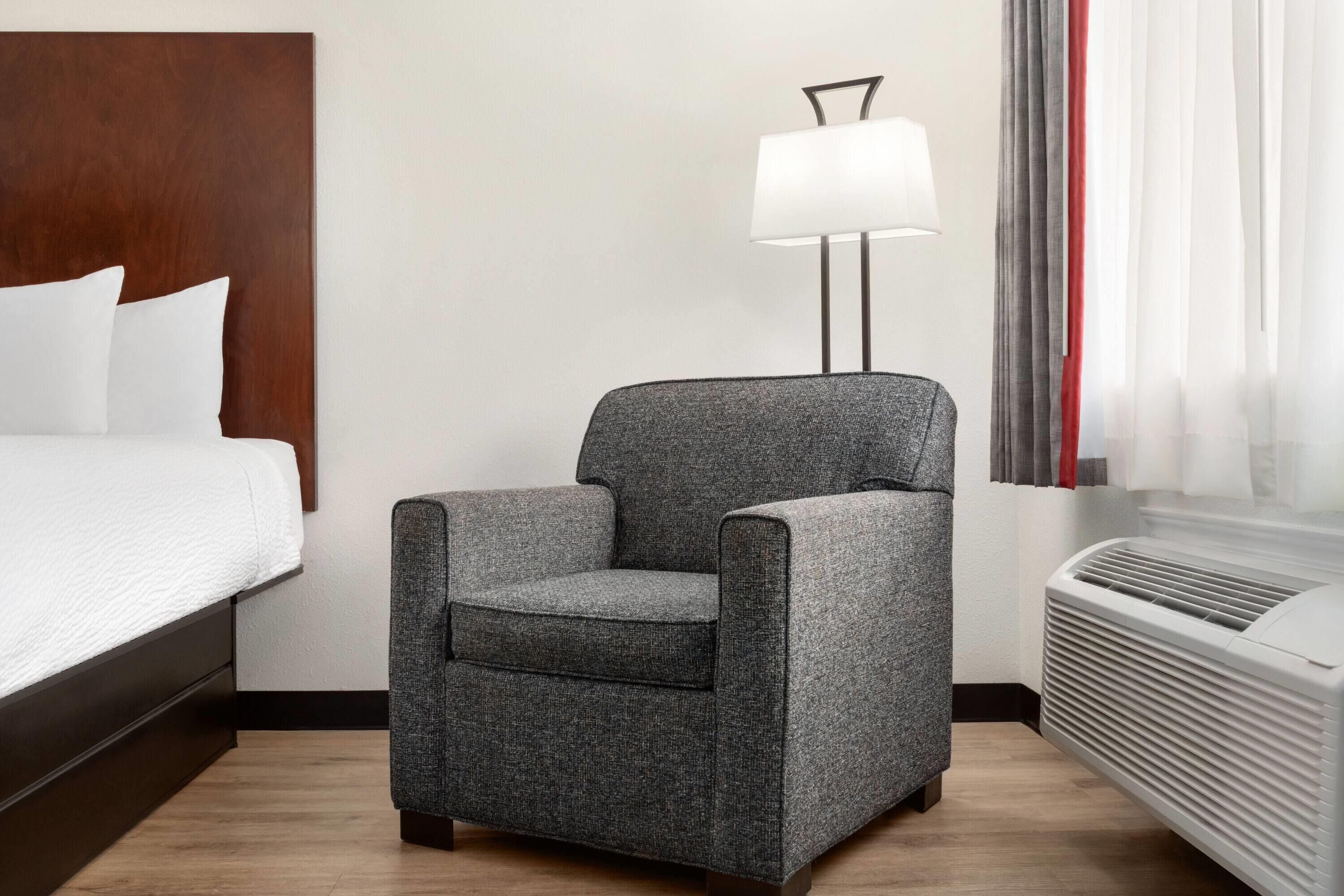 A hotel room with a bed, a grey armchair, a standing lamp, and a window with curtains.