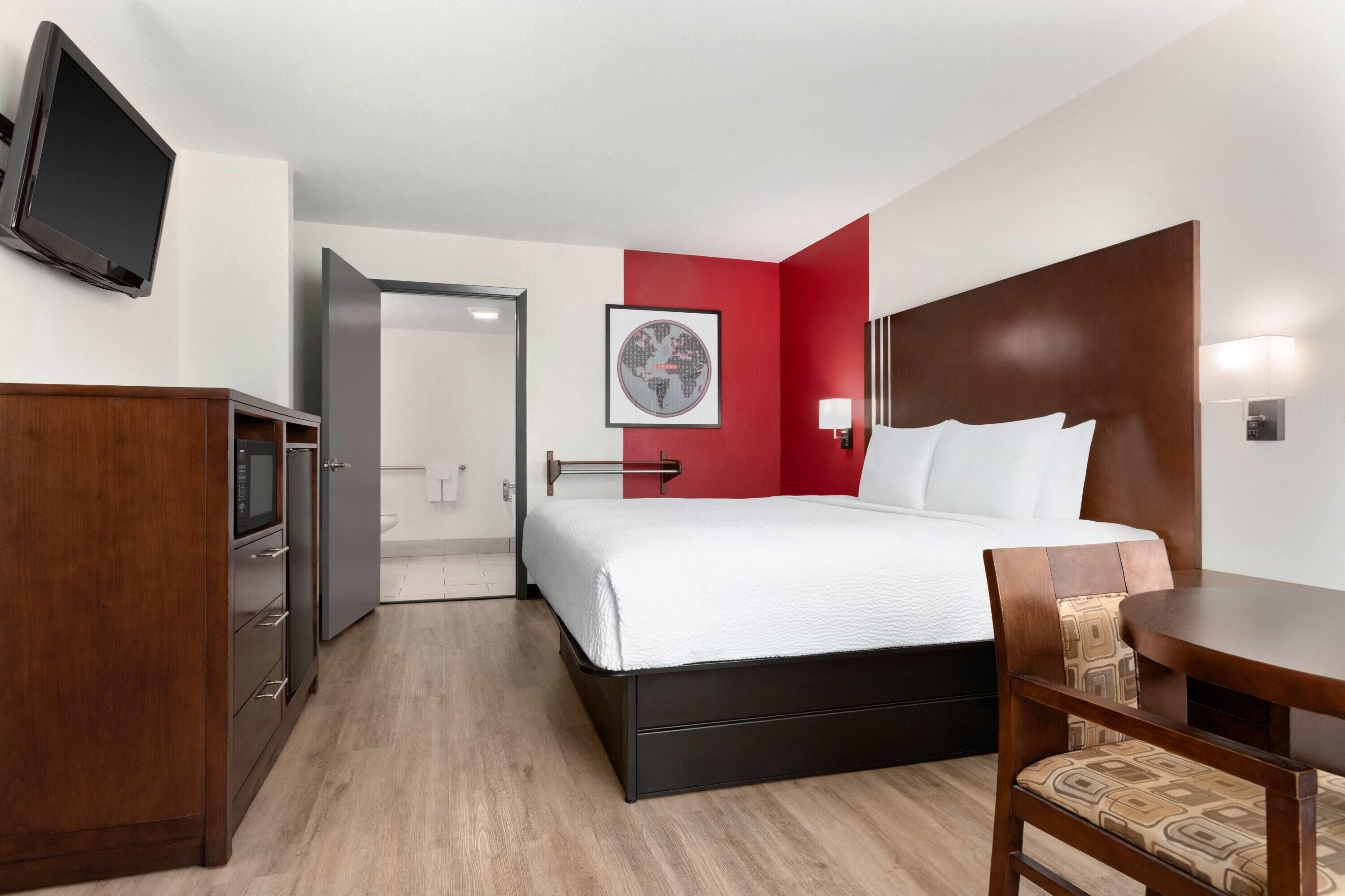 A hotel room with a bed, a desk, a chair, a TV, and a door leading to a bathroom.