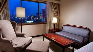 Une chambre d’hôtel avec vue sur la ville la nuit, un canapé, un fauteuil, une table et une lampe.