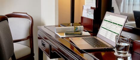 Quarto de hotel com escrivaninha de madeira, um laptop, um copo de água, luminária e uma cadeira.