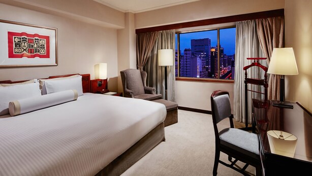 Une chambre d’hôtel avec un grand lit, un bureau avec une chaise, une lampe et une vue sur le paysage urbain au crépuscule.