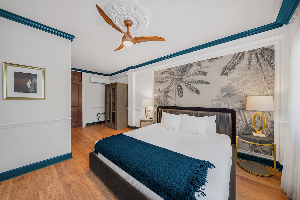 A hotel room with a large bed, a ceiling fan, a wall mural, and a wooden floor.