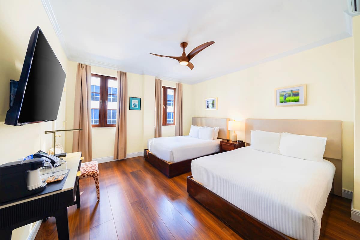 A hotel room with two beds, a desk with a telephone and a lamp, a ceiling fan, and a TV mounted on the wall.