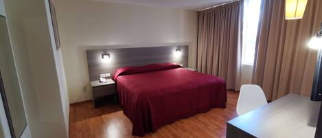 Habitación de hotel con piso de madera, cama con colcha roja, mesita de noche con teléfono, escritorio con lámpara y ventana con cortinas.