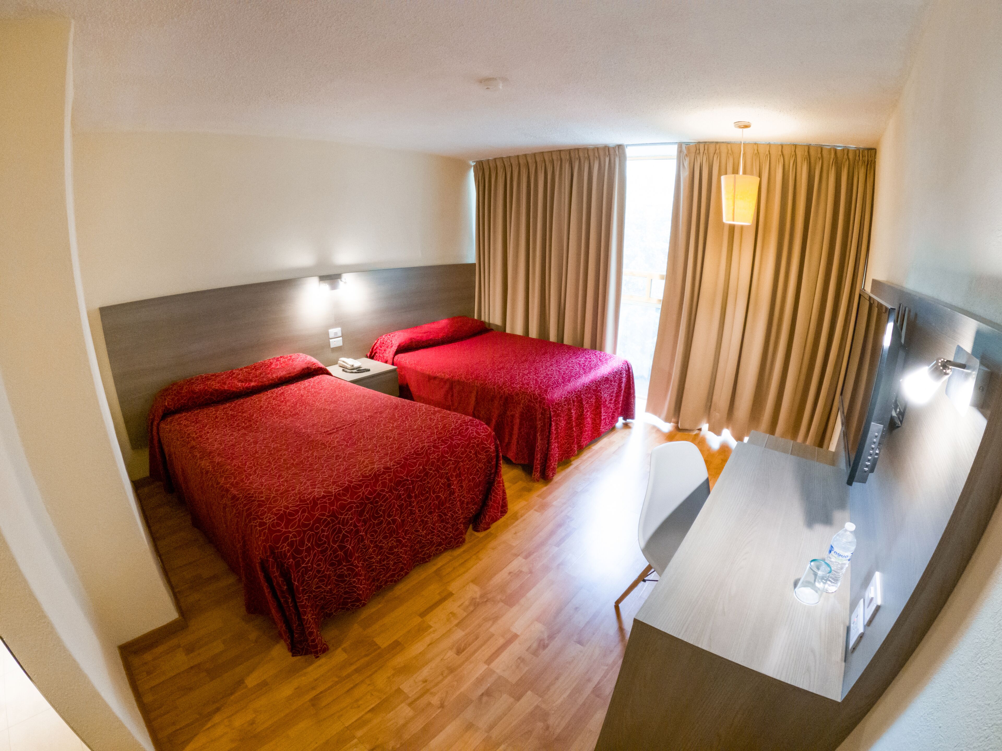 A hotel room with two single beds, a desk, a chair, a nightstand, and a window with curtains.