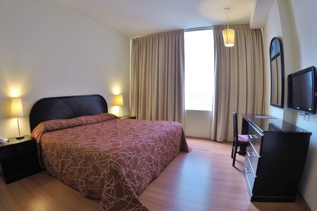 A hotel room with a large bed, a desk with a chair, a TV, and a window with curtains.