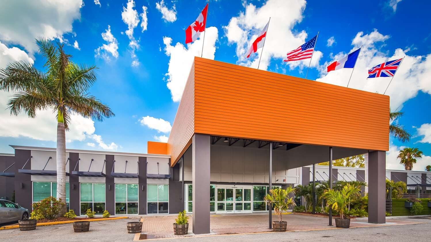 Un edificio moderno con un acceso cubierto, varias banderas y una palmera.