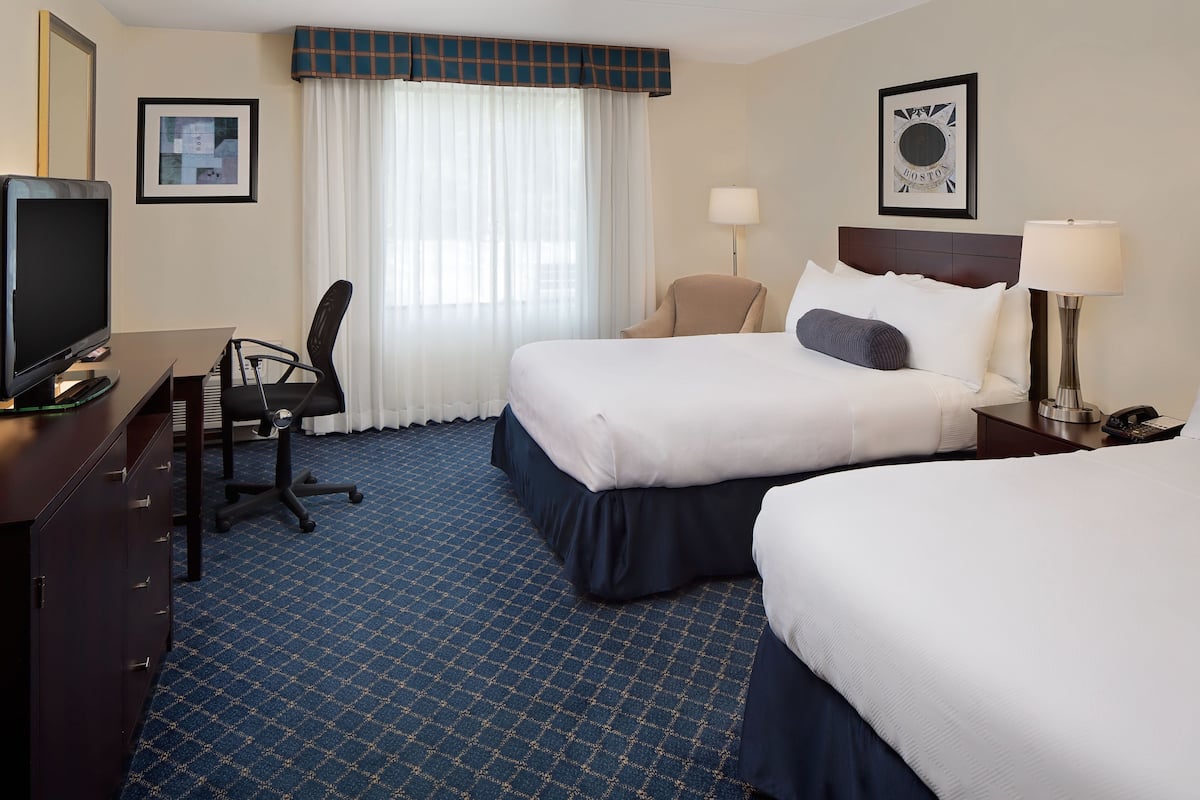 A hotel room with two beds, a desk with a television, a chair, a lamp, and a framed picture on the wall.