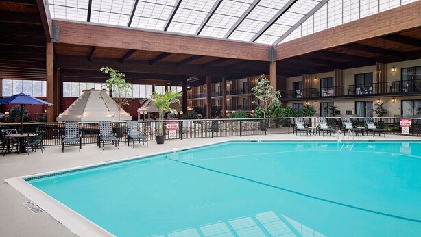 An indoor swimming pool with a glass ceiling, surrounded by lounge chairs and tables, and a staircase leading to upper levels.