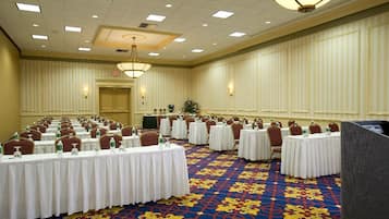 A conference room with multiple round tables, each with a white tablecloth and chairs. The room has a patterned carpet and a chandelier.