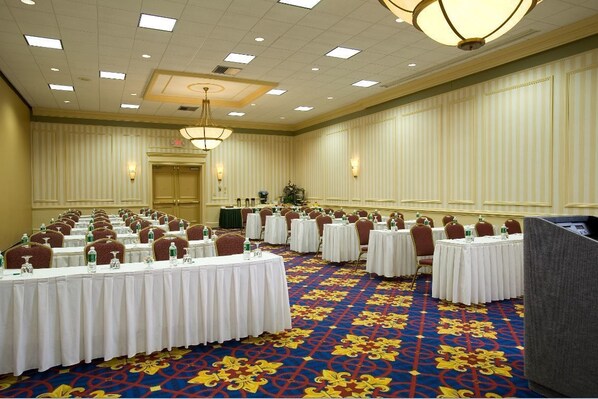 A conference room with multiple round tables, each with a white tablecloth and chairs. The room has a patterned carpet and a chandelier.