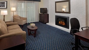 A hotel room with a sofa, armchair, small round table with a vase, a flat-screen TV on a cabinet, a fireplace, and a large mirror on the wall.