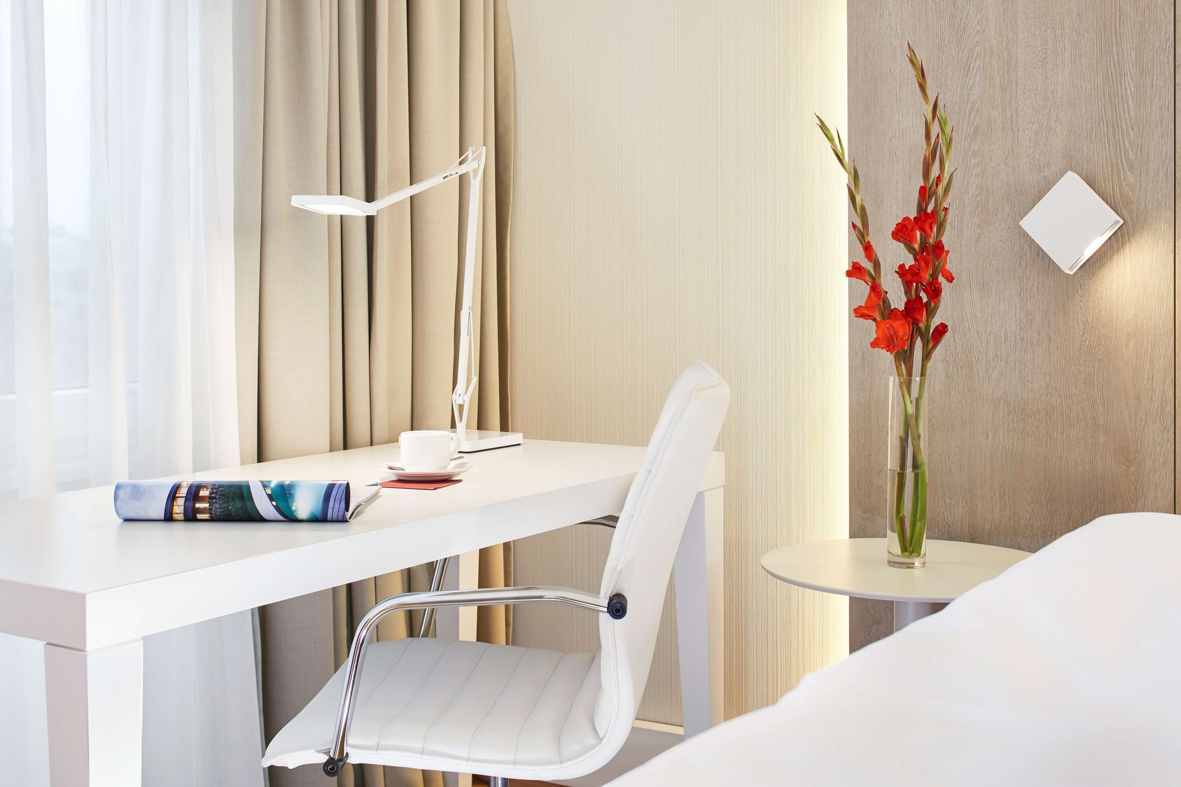 A hotel room with a white desk, a white chair, a bedside table with a vase of red flowers, and a lamp.