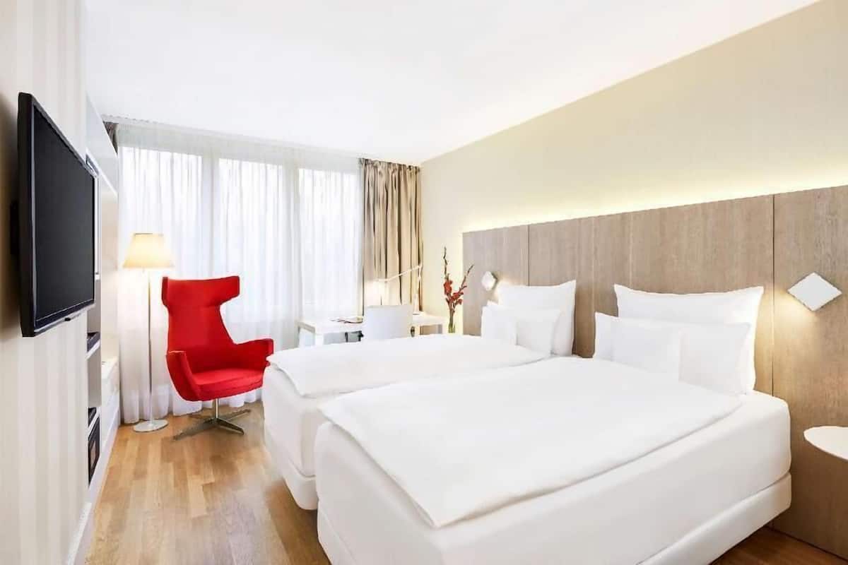 A hotel room with two beds, a red chair, a TV, and a desk with a lamp.