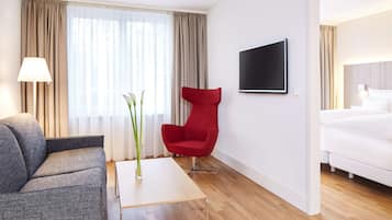 A modern hotel room with a grey sofa, a red armchair, a wooden coffee table with a vase, a bed, and a TV mounted on the wall.