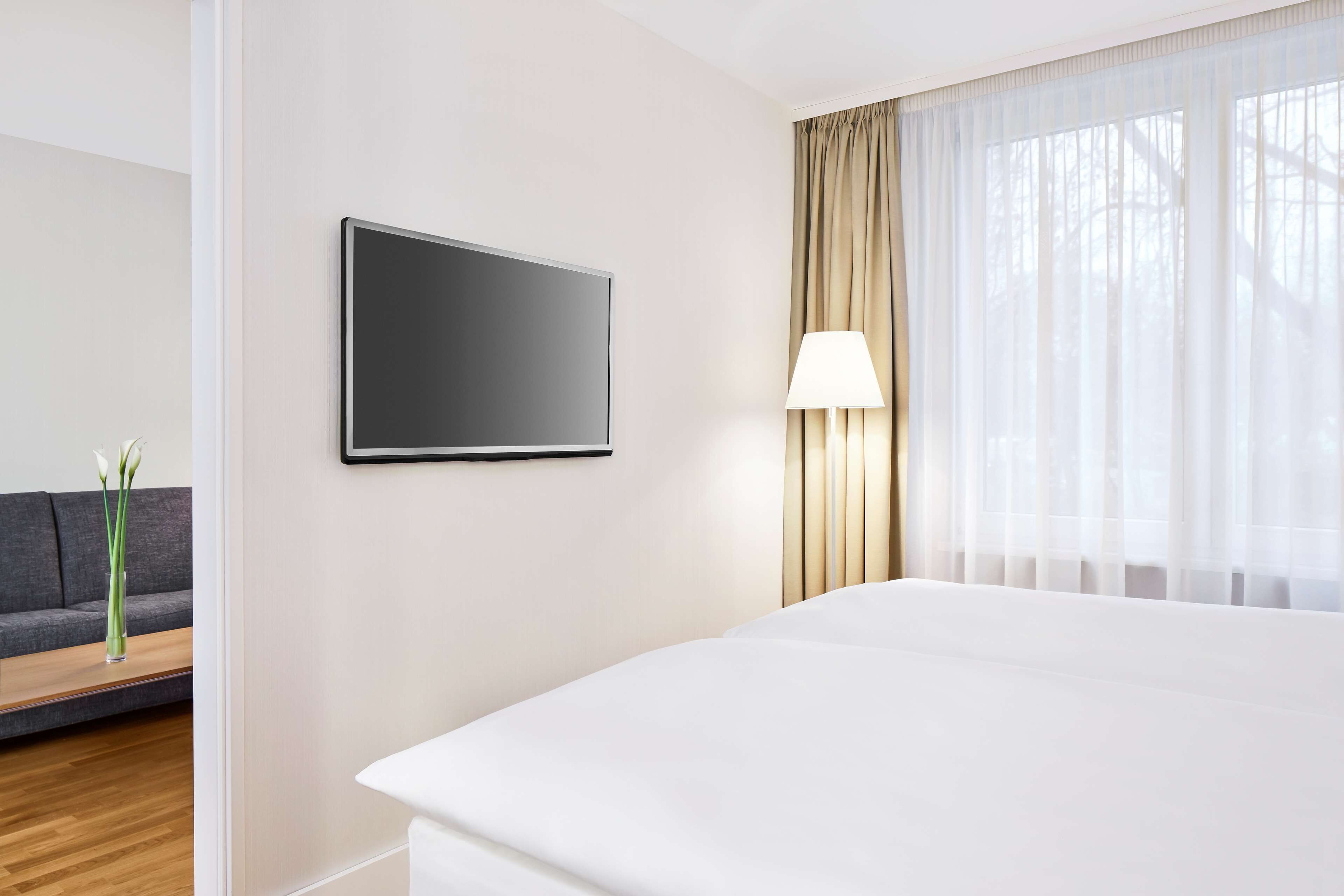 Ein Hotelzimmer mit einem großen Bett, einem an der Wand montierten Flachbildfernseher, einer Nachttischlampe und einem Fenster mit durchsichtigen Vorhängen.
