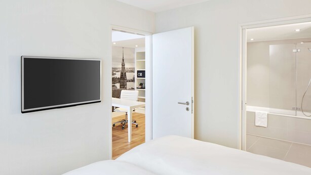 A modern hotel room with a bed, a TV mounted on the wall, a shower area, and a desk with a chair. - NH Collection Hamburg City (Hamburg)