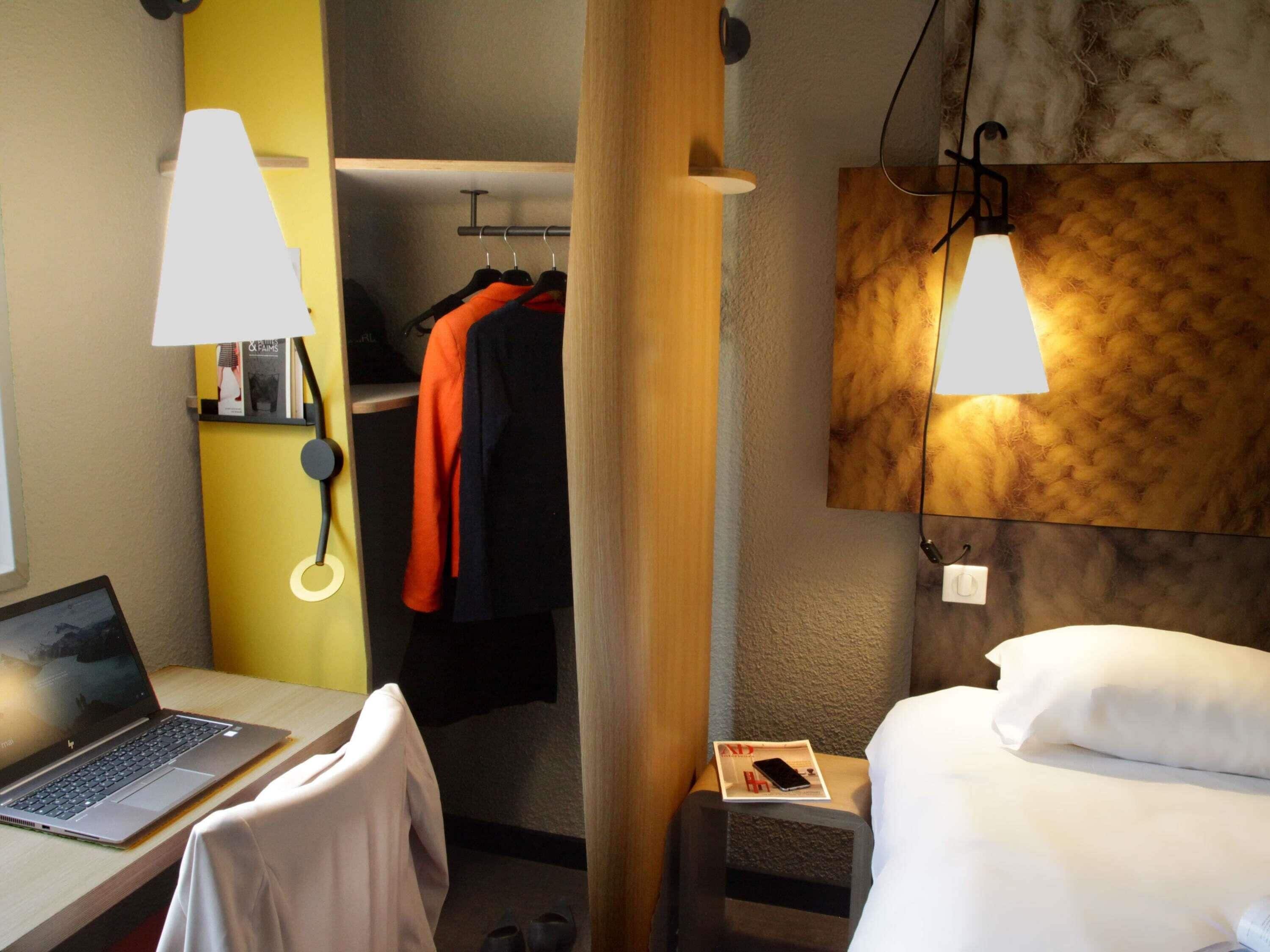 A hotel room with a bed, a desk with a laptop, a closet with hanging clothes, and a wall-mounted lamp.