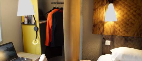 A hotel room with a bed, a desk with a laptop, a closet with hanging clothes, and a wall-mounted lamp.
