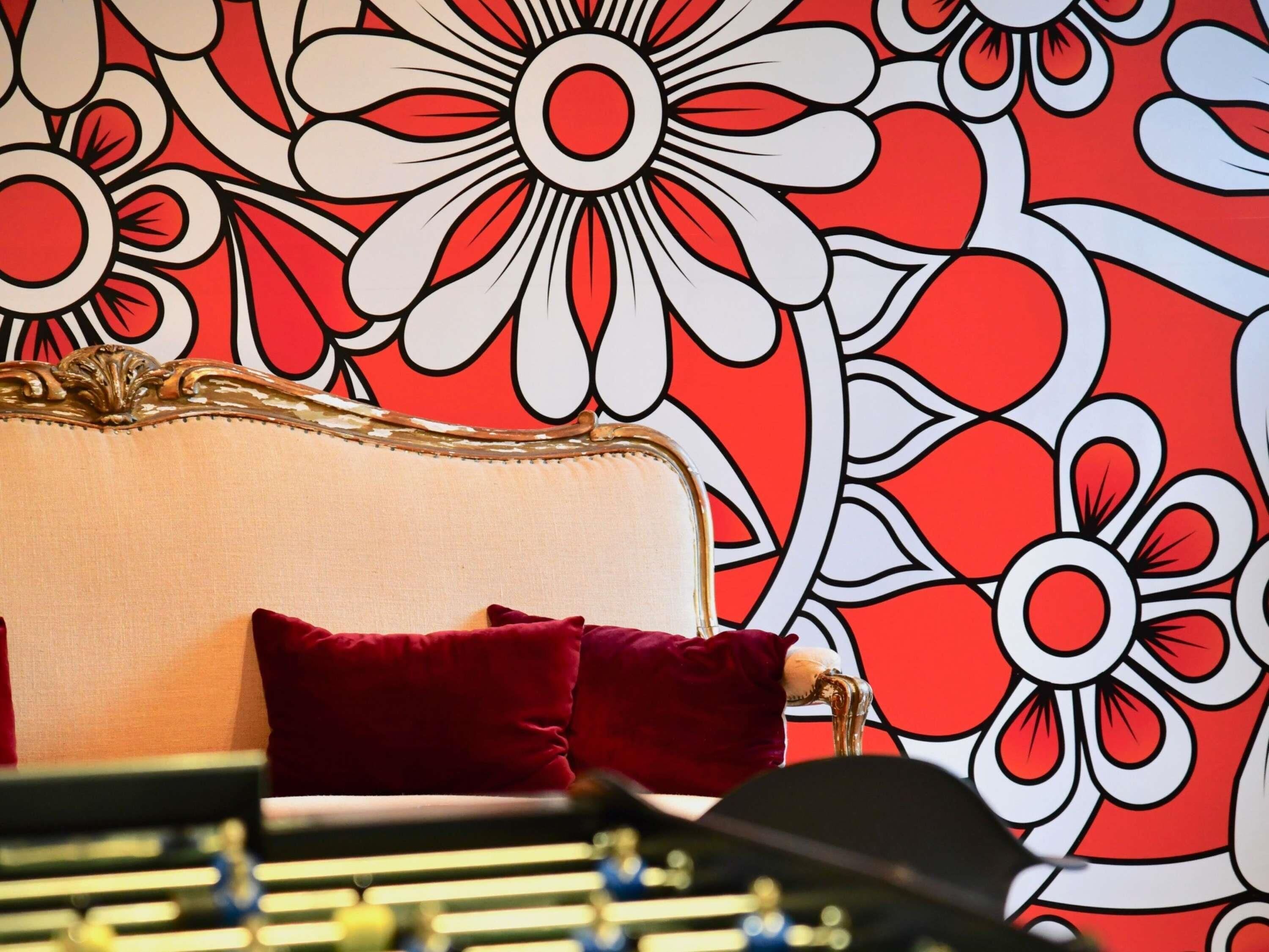 A room with a patterned wall, a bed with red pillows, and a gold-framed headboard.