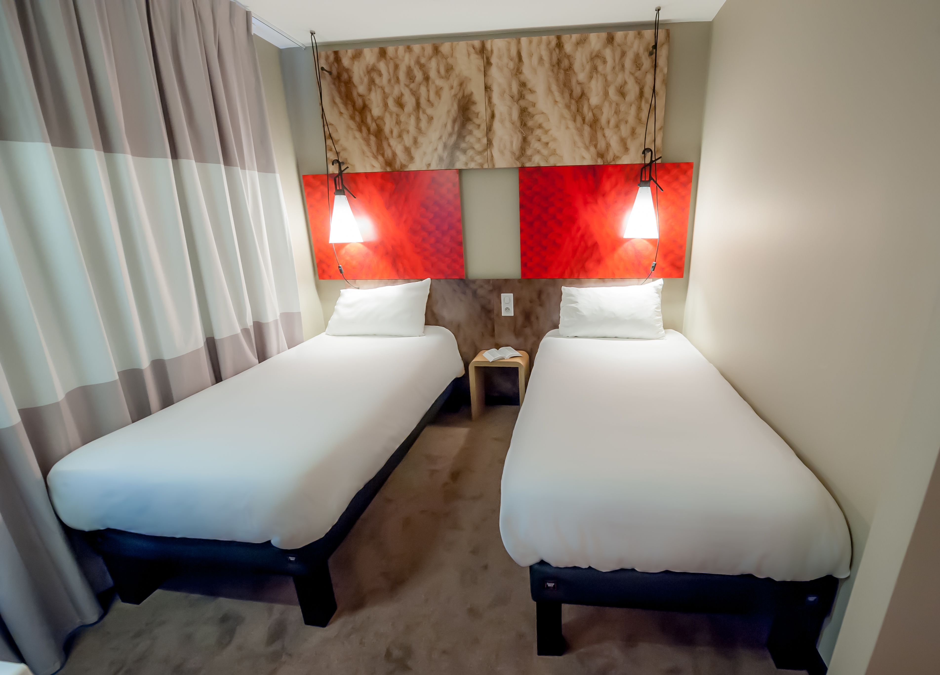A hotel room with two single beds, a nightstand with a book, and a red and beige textured wall.