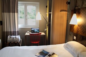 A hotel room with a bed, a desk with a laptop, a red chair, a white bag, and a window with curtains.