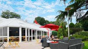An outdoor seating area with tables and chairs, a covered section with large windows, and a red umbrella providing shade. - ibis Quimper (Quimper)