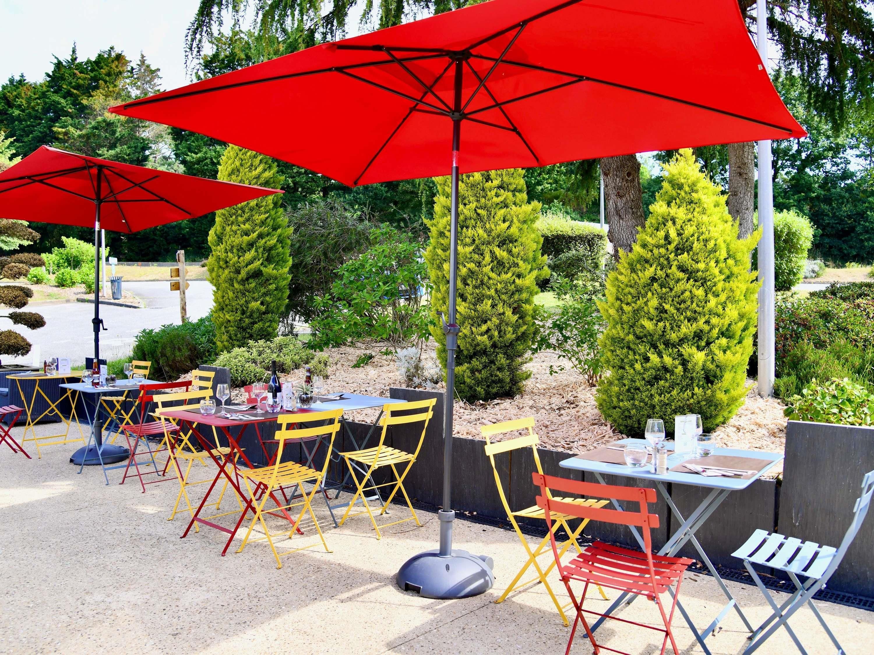 Outdoor tables with red umbrellas, yellow and red chairs, and a landscaped garden in the background.