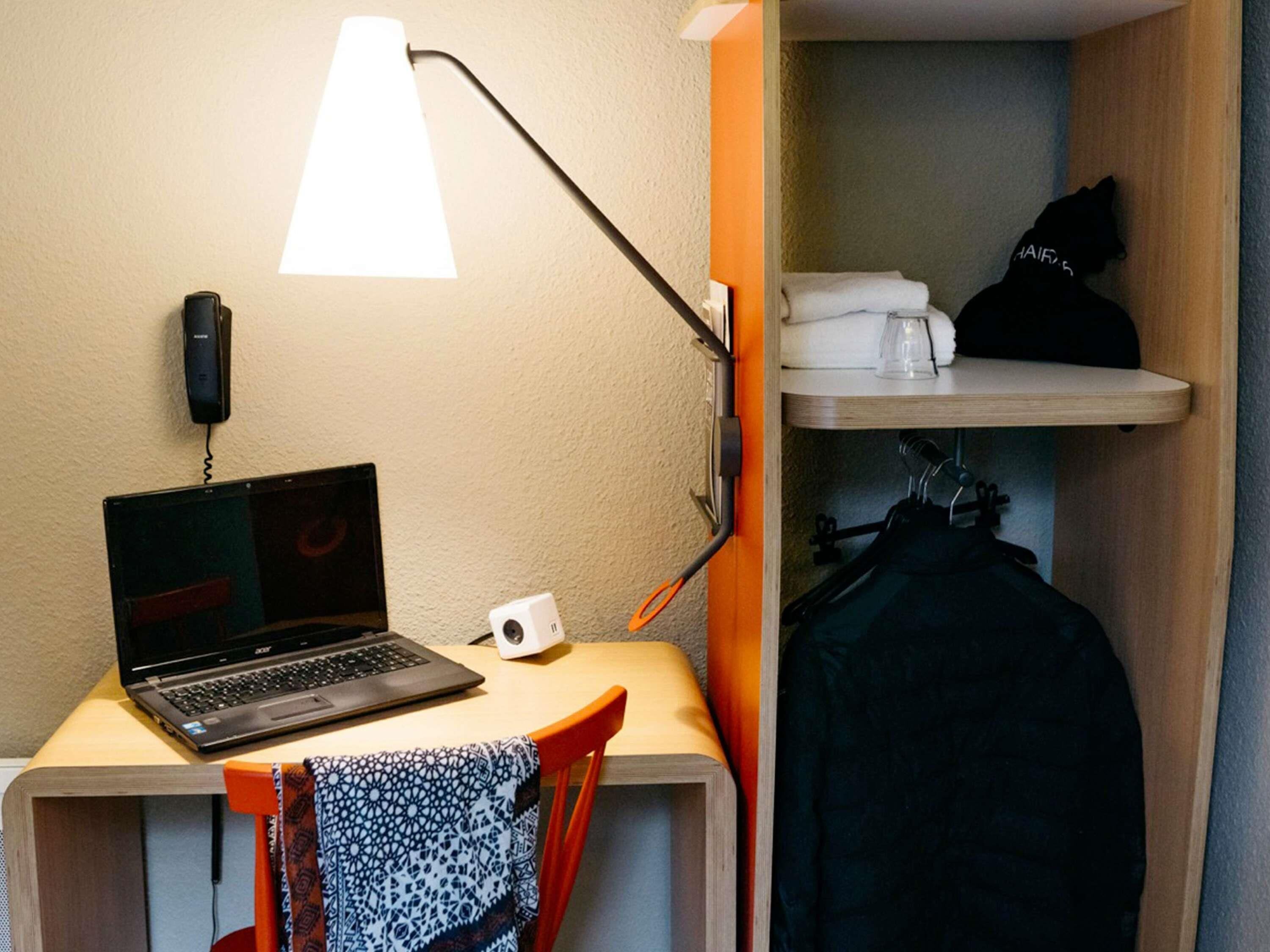 A laptop on a desk with a lamp, a phone, and a cup. There is a chair with a patterned cloth and a coat rack with a jacket.