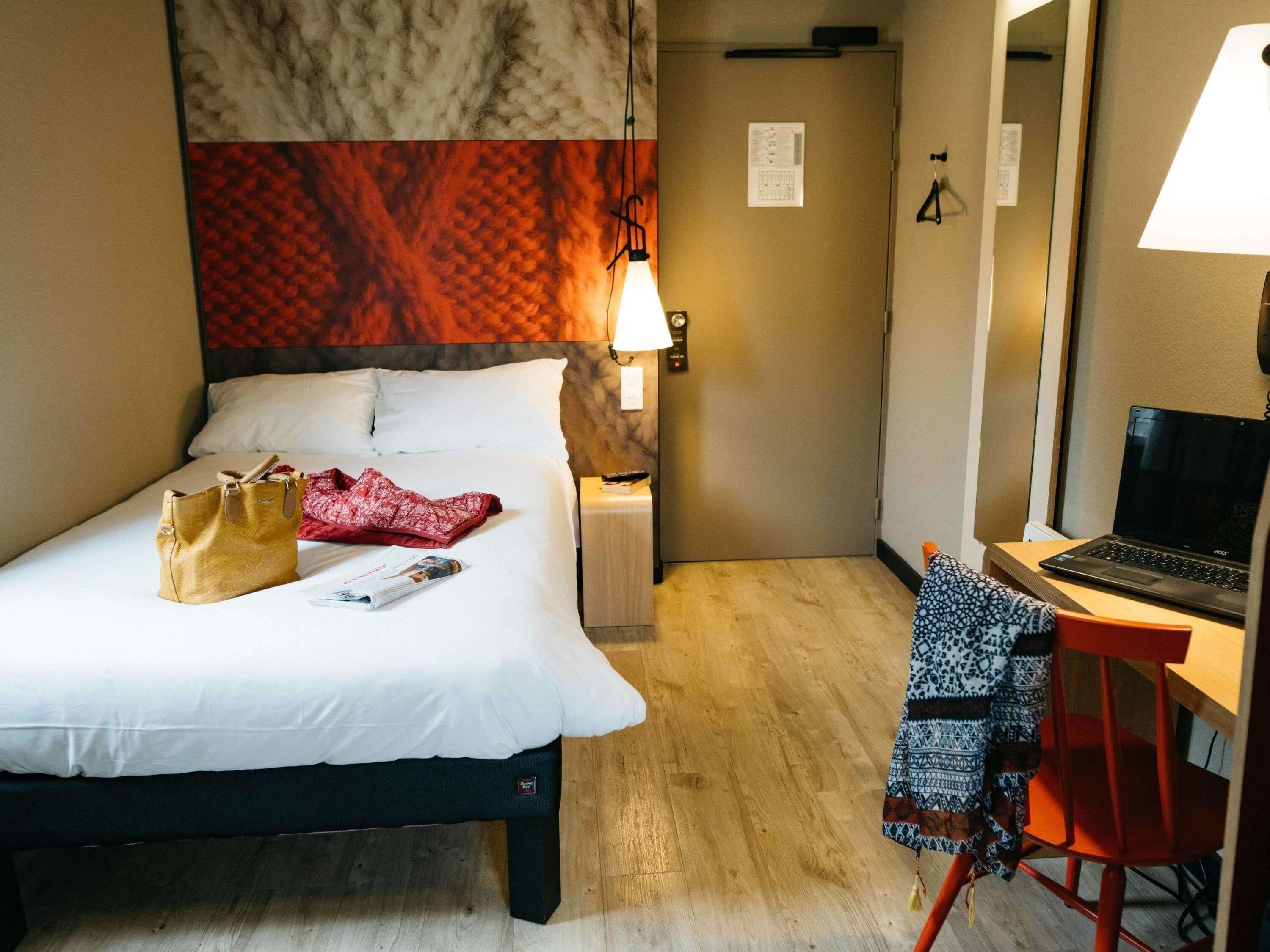 A hotel room with a bed, a desk with a laptop, a chair, a lamp, and a door with a sign.