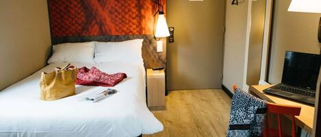 A hotel room with a bed, a desk with a laptop, a chair, a lamp, and a door with a sign.