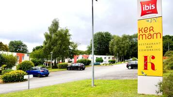A sign for 'ibis Hotels' and 'ker mam restaurant bar coworking' with a road and parked cars in the background.