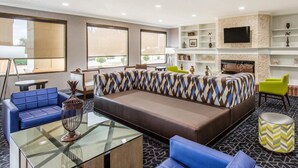 A modern lounge area with a sectional sofa, blue armchairs, a glass coffee table, and a fireplace. - Comfort Inn College Park North (Beltsville)
