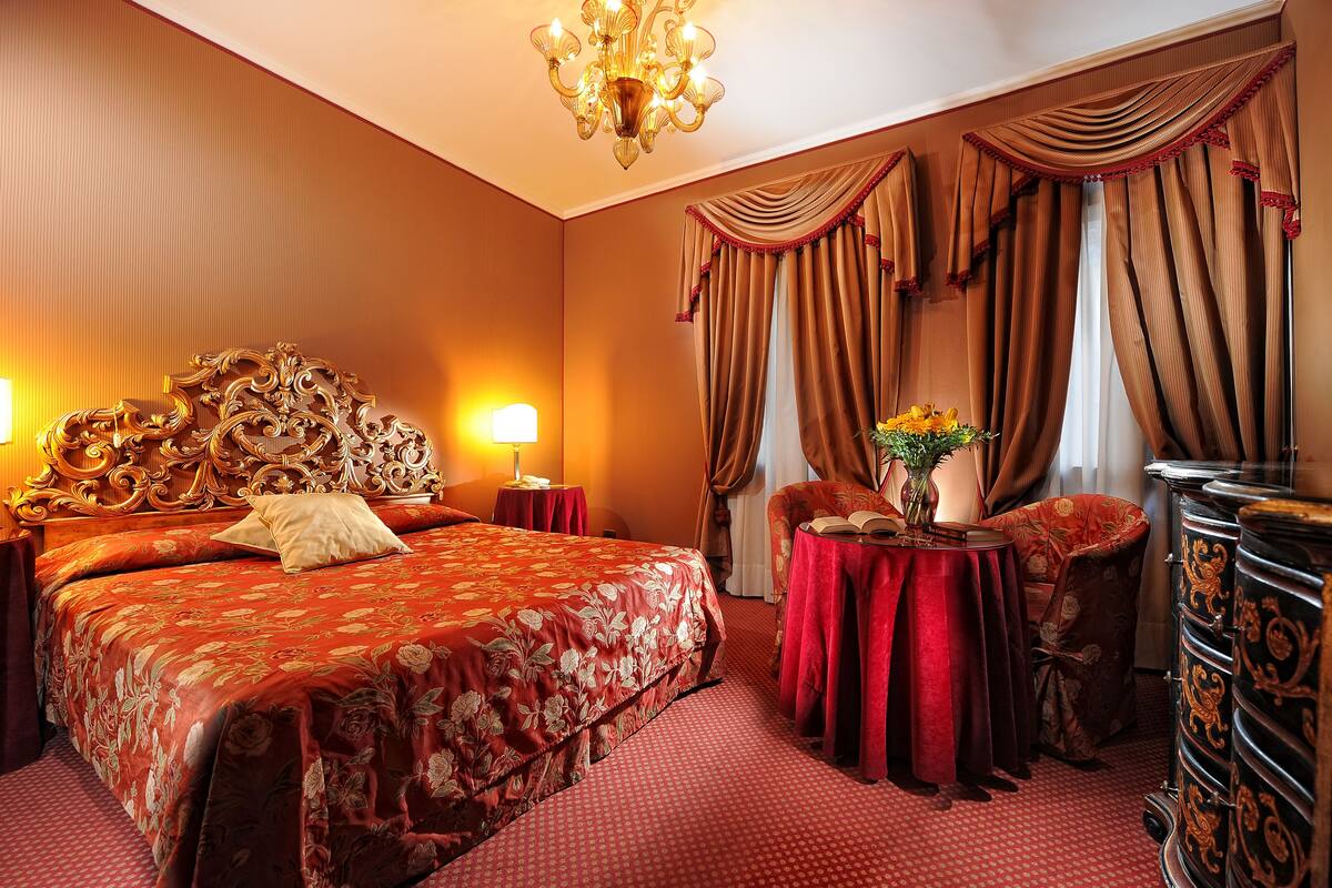 A hotel room with a large bed, a chandelier, a desk with a vase, and a dresser.