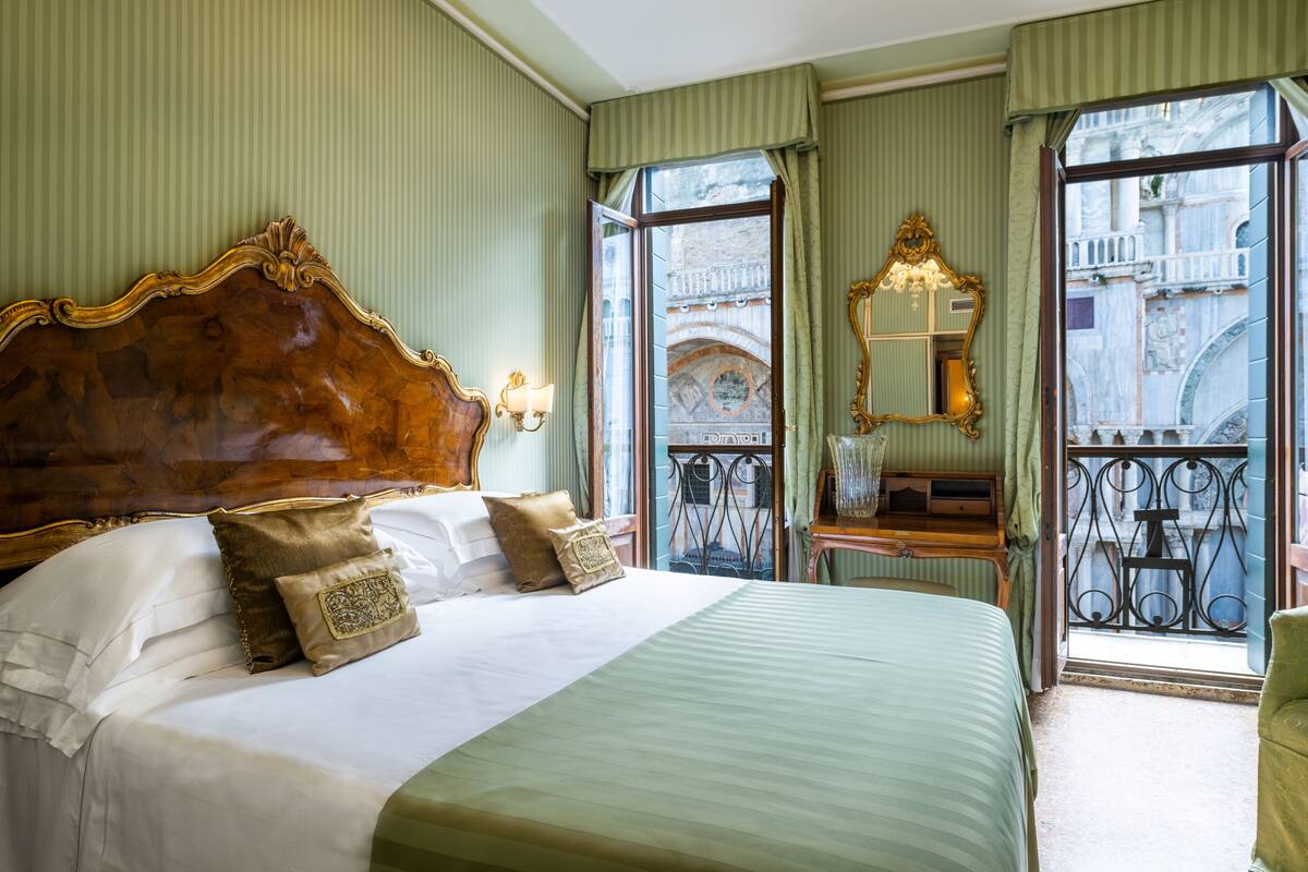 A hotel room with a large bed, a small table with a mirror, and a balcony with a wrought iron railing.