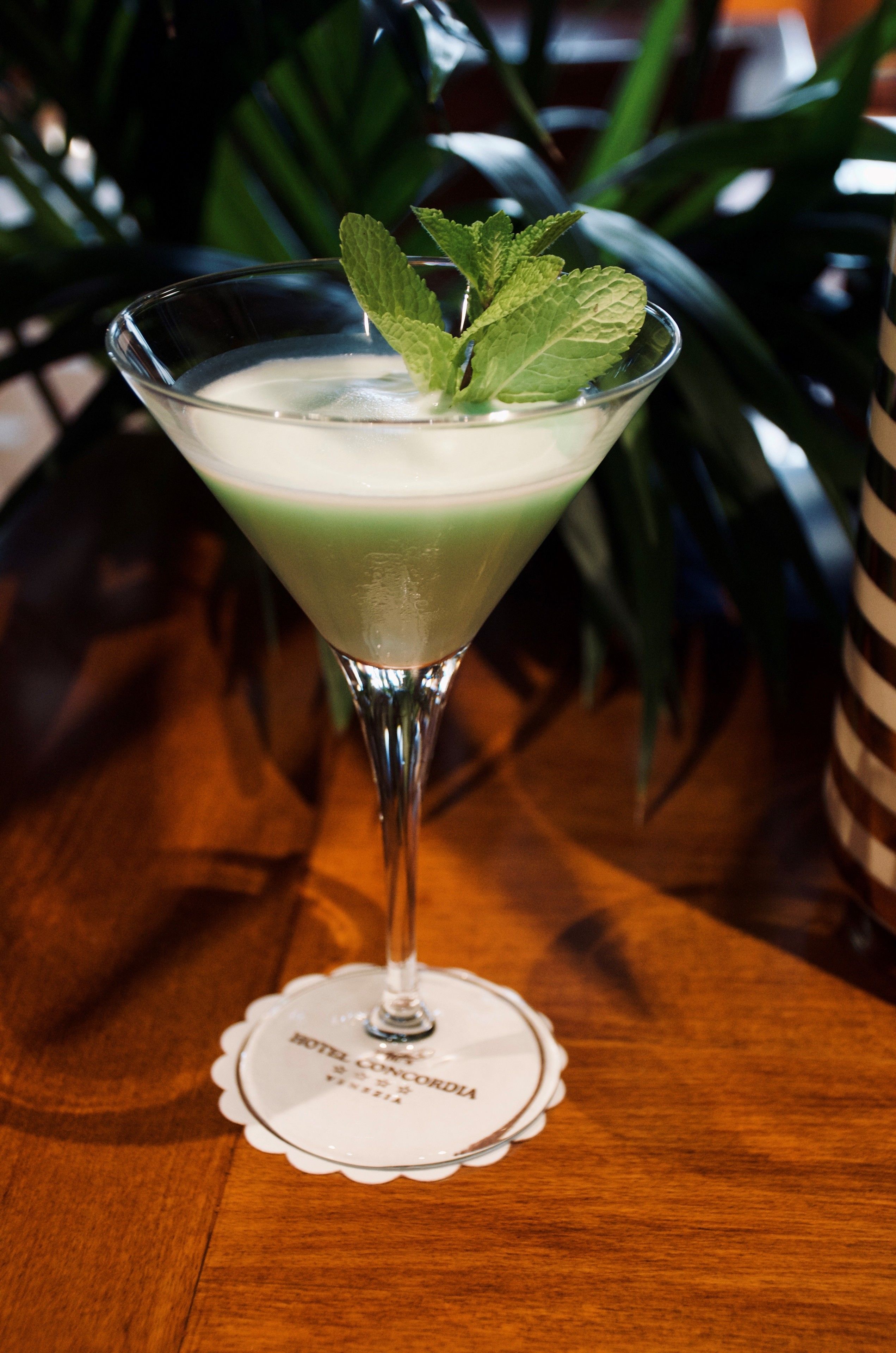 A martini glass with a green drink, garnished with a mint leaf, on a wooden table with a placemat.