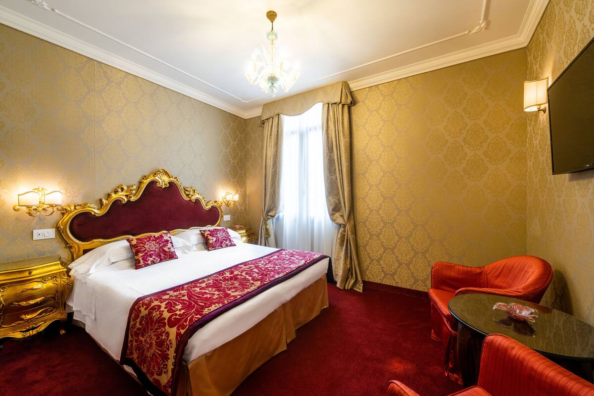 A hotel room with a large bed, a chandelier, a television, and red upholstered chairs.