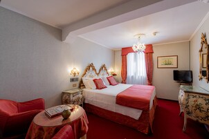 Ein Hotelzimmer mit einem großen Bett, rot-weißer Bettwäsche, einer Kandelaber, einem Fernseher und einem kleinen Tisch mit einer Vase.