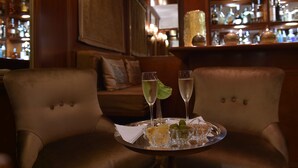 Una zona de bar con dos sillas, una bandeja con vasos y aperitivos, y una estantería con botellas.
