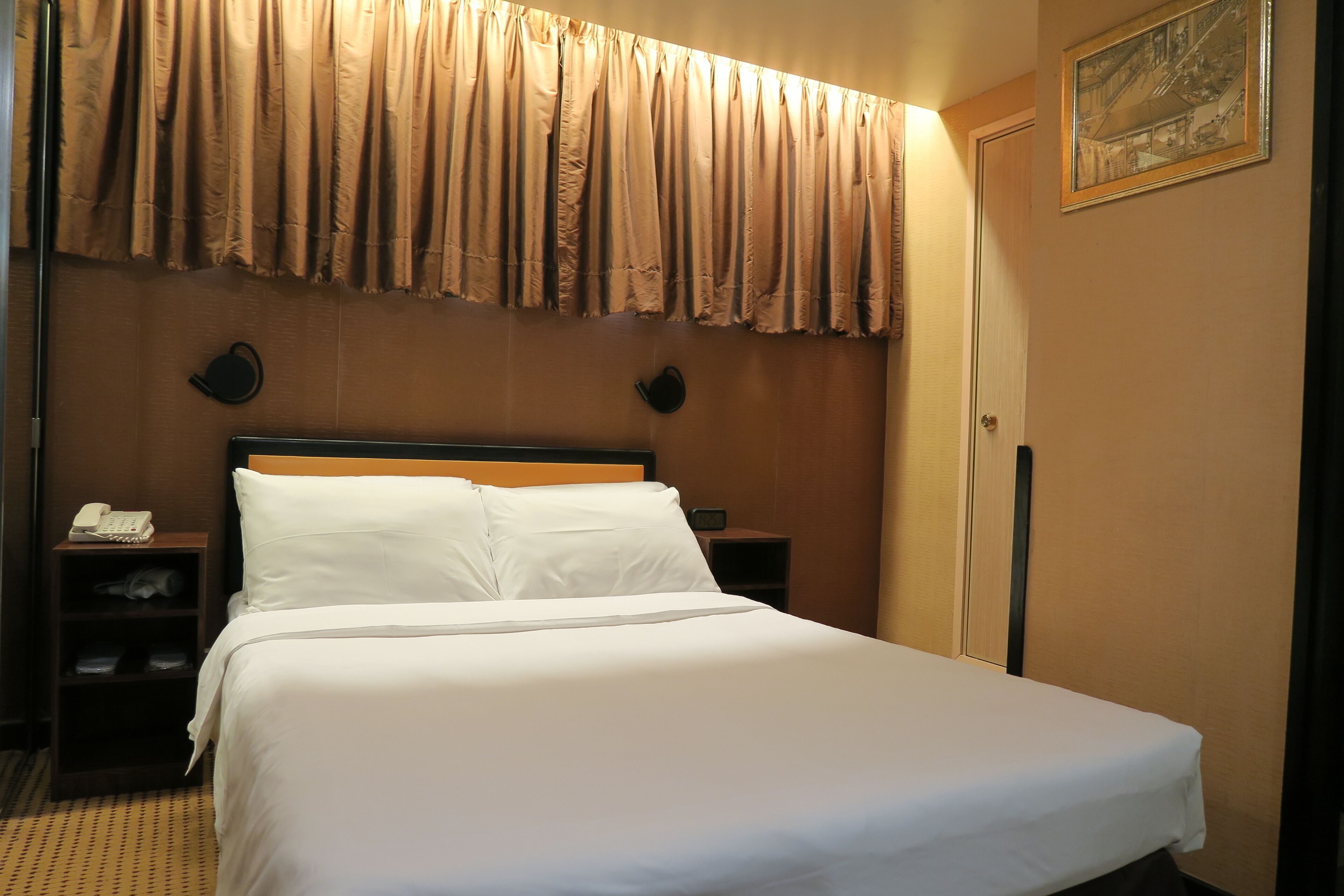 A hotel room with a large bed, a nightstand with a telephone, and a wall-mounted clock.